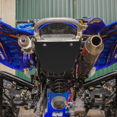 Traseira de quad azul com pneus grandes, escape metálico e suspensão visível num espaço externo