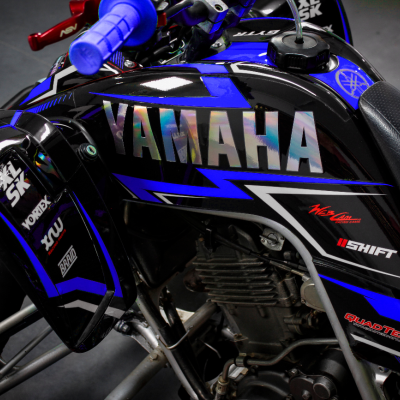Quadro azul e preto de mota Yamaha com texto YAMAHA refletivo no depósito e várias outras inscrições coloridas.