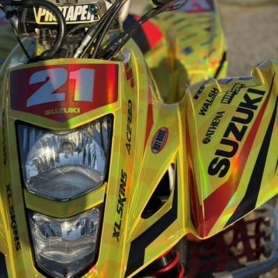Quadriciclo amarelo Suzuki com número 21, farol duplo e vários adesivos de marcas