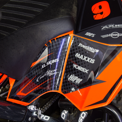 Assento preto de moto com traseira laranja e vários logotipos em ambiente de piso escuro