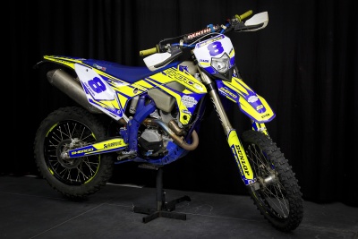 Motocross Sherco azul e amarelo com número 8 e logos visíveis em fundo preto.