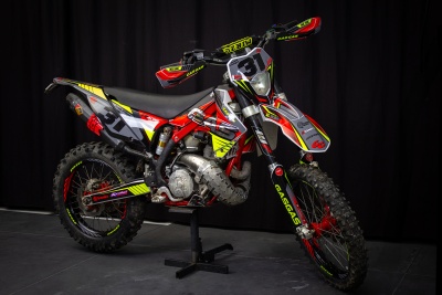 Motocross vermelha, preta, amarelo e branca com grafismos, apoiada em suporte em fundo escuro