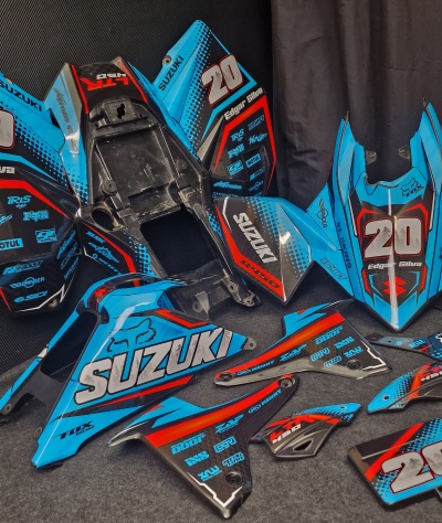 Carenagens de mota Suzuki azuis com detalhes vermelhos e pretos, texto 'SUZUKI', '20', 'Edgar Silva', várias marcas e logotipos, sobre chão cinzento