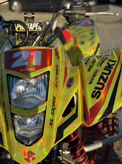 Quadriciclo amarelo Suzuki com número 21, farol duplo e vários adesivos de marcas