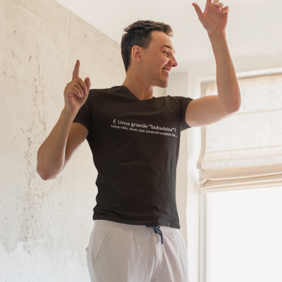 Homem com camiseta preta com texto branco e calças cinza claro em interior com parede branca e janela com estore bege.