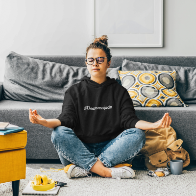 Pessoa meditando com sweatshirt preta '#Deusmaajude', jeans, óculos, sentada num tapete cinzento, com acessórios perto e sofá ao fundo
