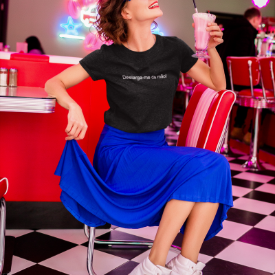 Mulher com camiseta preta, saia azul e ténis brancos sentada em diner retro com copo de batido rosa.
