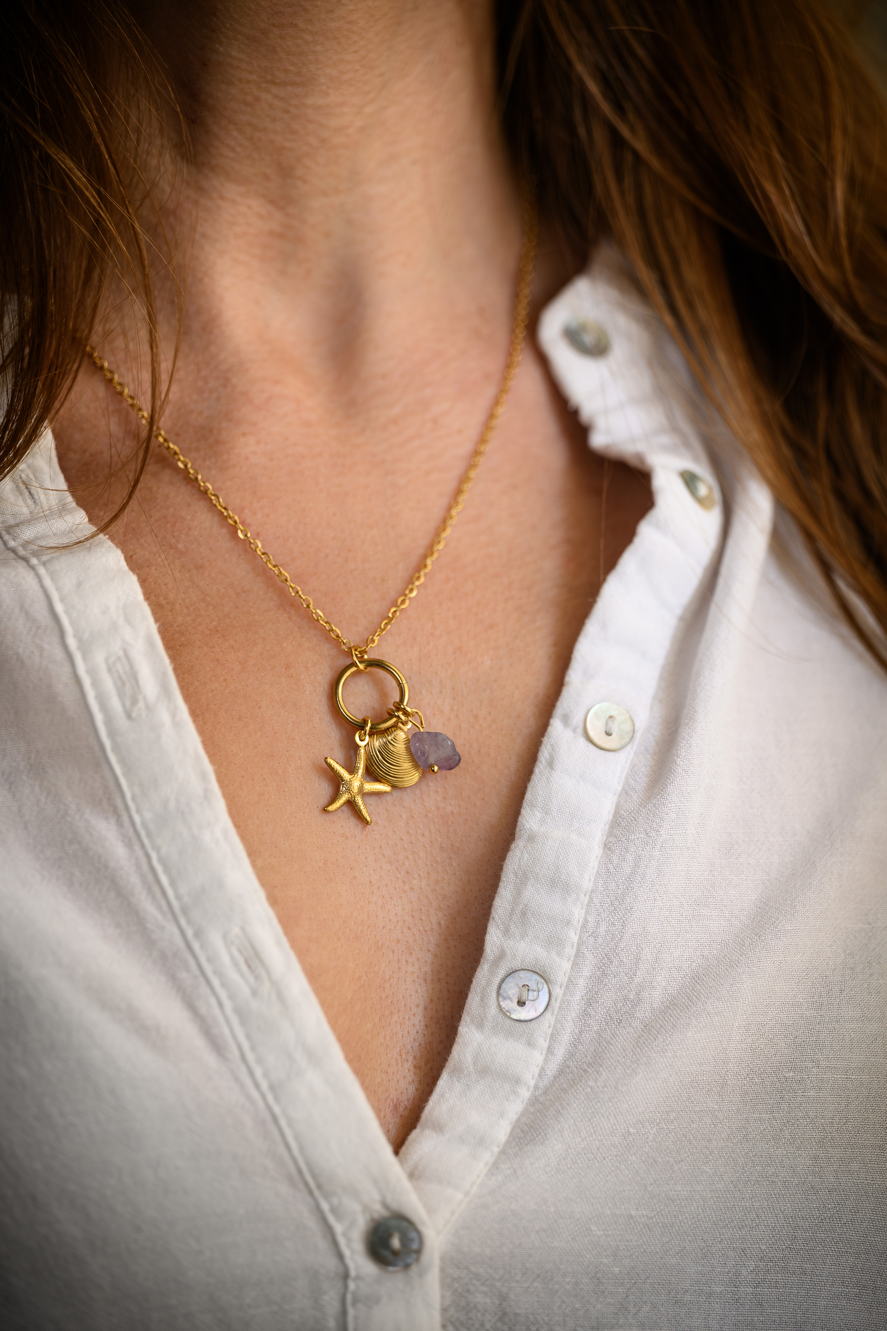 Colar dourado com pingentes de estrela-do-mar, concha e pedra roxa ao pescoço, camisa branca aberta