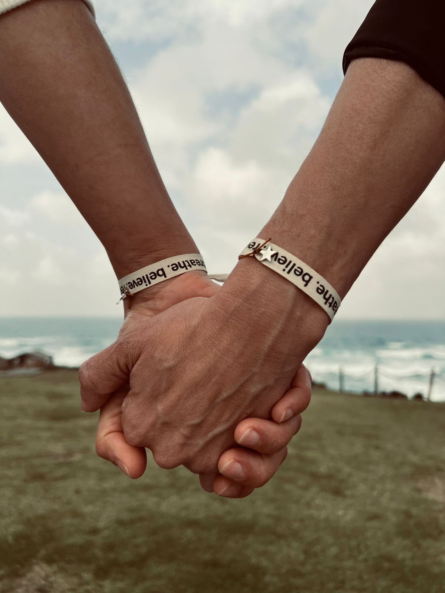 Duas mãos dadas com pulseiras têxteis BeThe frente ao mar e céu nublado