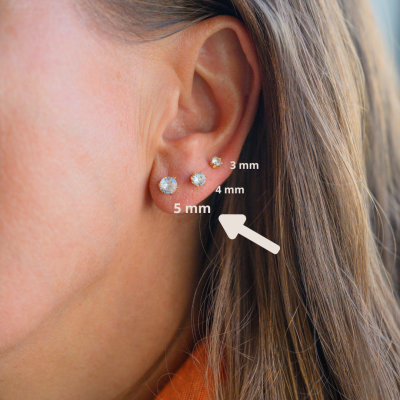 Três brincos redondos brilhantes em orelha feminina com tamanhos indicados.