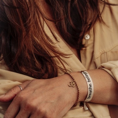 Pulseiras e anéis em braço com camisa bege, destaque para texto breathe_ believe na pulseira de tecido