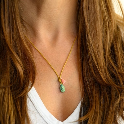 Colar com pendente verde e rosa em cordão amarelo, usado por pessoa com cabelo castanho claro e camisa branca