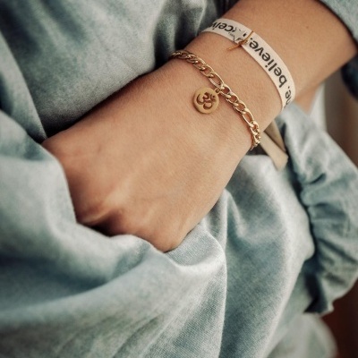 Pulseira dourada com pendente símbolo OM no pulso de pessoa com camisa cinzenta