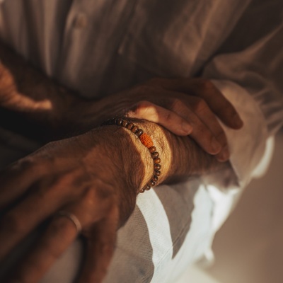 Pulseira de contas castanhas com conta amarela numa mão masculina cruzada sobre a outra, em roupa clara