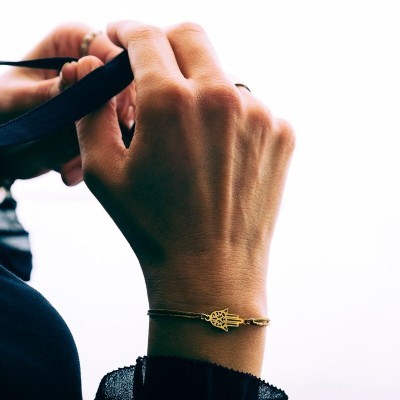 Pulseira dourada com pendente em forma de mão no pulso de uma pessoa com roupa preta