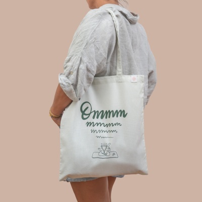 Saco de tecido branco com texto verde e desenho de gato sentado num tapete de ioga.