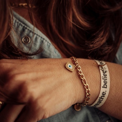 Pulseira dourada com pendente olho azul e pulseira branca com texto no pulso