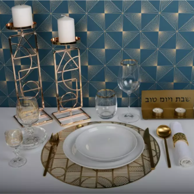 Mesa posta com pratos brancos, talheres dourados, copos, castiçais com velas e placa com texto em hebraico