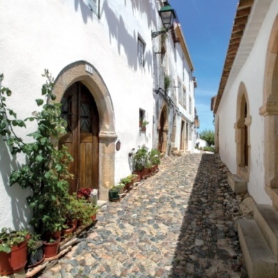 Rua estreita com calçada portuguesa e edifícios brancos sob céu azul
