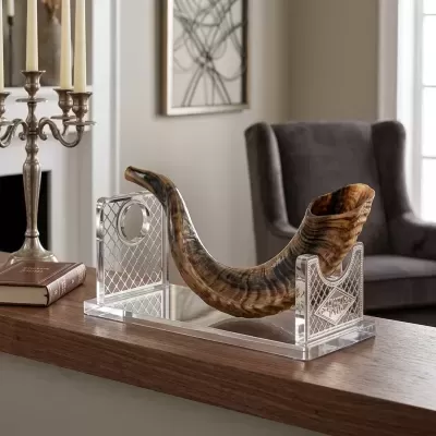 Corneta de chifre castanha e preta num suporte transparente sobre mesa de madeira numa sala com braço de cadeira cinza, castiçal e livro.