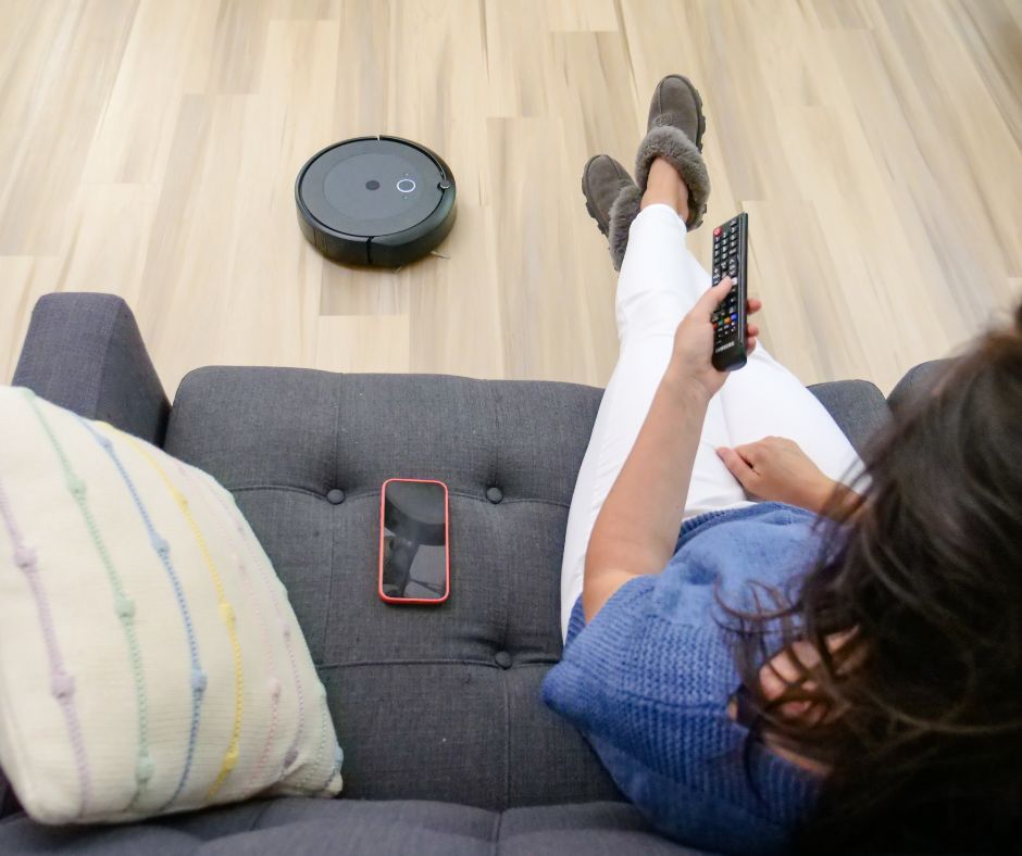 Robô aspirador sobre chão de madeira ao lado de sofá cinzento com pessoa e telemóvel.