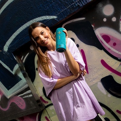 Mulher com vestido rosa claro segurando coluna sonora azul em frente a graffiti