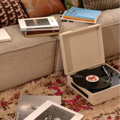 Gira-discos branco aberto a tocar vinil ao lado de discos e sofá com almofadas coloridas