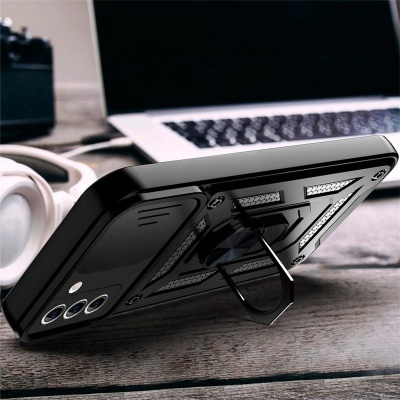 Capa de telemóvel preta com suporte metálico e textura geométrica sobre uma mesa com portátil e auscultadores ao fundo