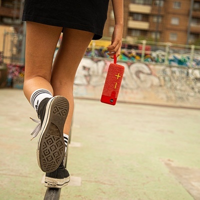 Coluna portátil vermelha com símbolos de mais e menos, mãos de pessoa com ténis, parque skate urbano.