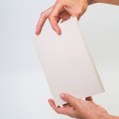 Bloco de notas creme com letras em relevo 'ASK' segurado por mãos