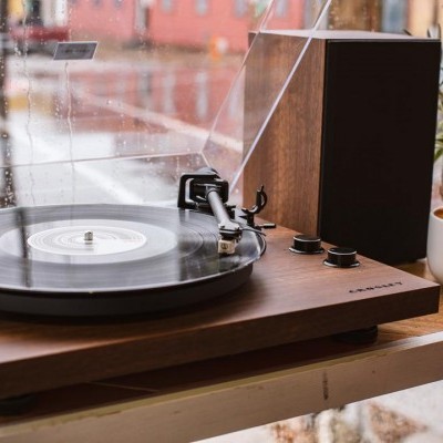 Gira-discos de madeira com prato preto e tampa transparente numa mesa perto de uma janela molhada pela chuva e um copo branco ao lado