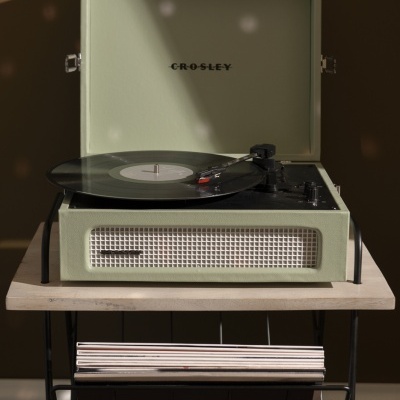 Gira-discos Crosley verde claro vintage com disco de vinil e rack com discos em fundo castanho