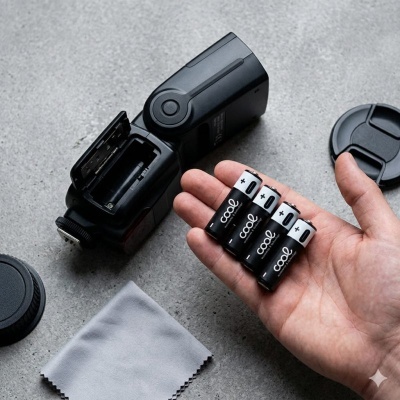 Mão segurando pilhas COOL com flash fotográfico e acessórios sobre superfície cinzenta