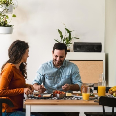 Casal a jantar numa mesa de madeira com comida e bebidas num ambiente com plantas e paredes brancas