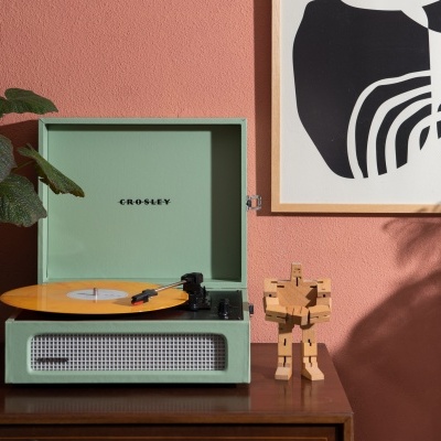 Gira-discos Crosley verde com disco vinil amarelo em móvel de madeira junto a figura de madeira e plantas