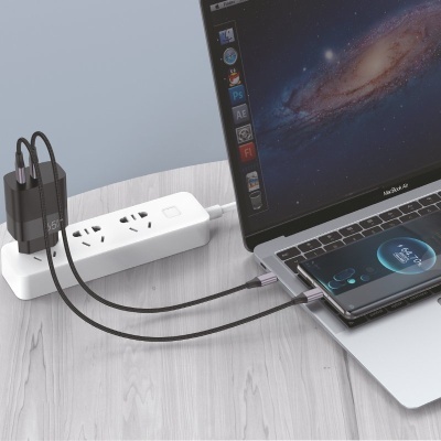 Cabo USB tipo C preto entre MacBook Air e smartphone, ligado a régua de tomadas branca numa mesa de madeira clara