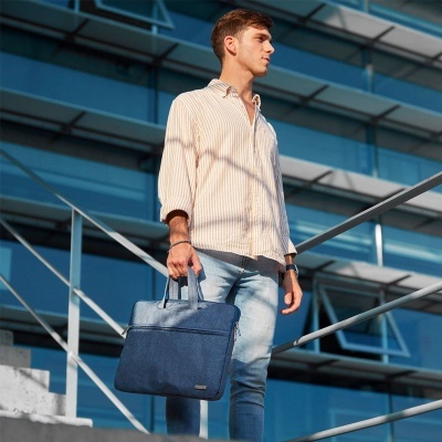 Homem jovem com camisa bege riscada, calças de ganga e mala azul escura em ambiente urbano