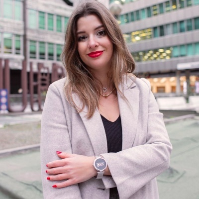 Mulher com relógio de pulso cinza e bracelete preta, casaco bege e blusa preta, em área urbana com edifícios modernos.