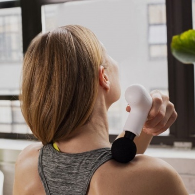 Mulher a usar massageador corporal portátil branco na zona do ombro