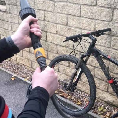 Homem a segurar mangueira de limpeza junto a bicicleta preta encostada em parede de pedra.