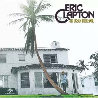 Capa de álbum com casa branca, palmeira inclinada e Eric Clapton em pé à frente