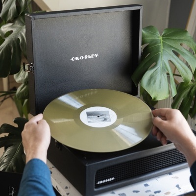 Gira-discos portátil preto CROSLEY com disco vinil dourado e plantas ao fundo