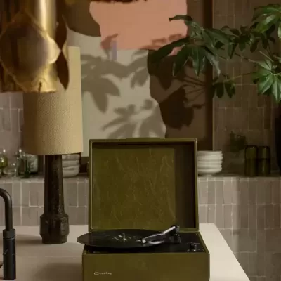 Toca-discos verde oliva da marca Crosley numa cozinha com azulejos bege e frutos numa taça