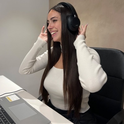 Mulher a usar auscultadores pretos, camisola branca, sentada numa cadeira preta junto a computador portátil