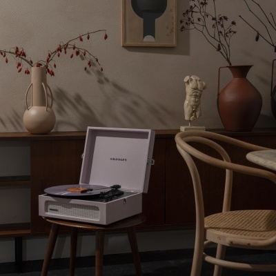 Toca-discos branco com disco roxo sobre mesa de madeira, cadeira clara e decoração com vasos e quadro abstrato