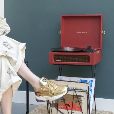 Gira-discos Crosley vermelho com discos de vinil num suporte metálico ao lado de uma pessoa com sapatilhas douradas e saia creme.