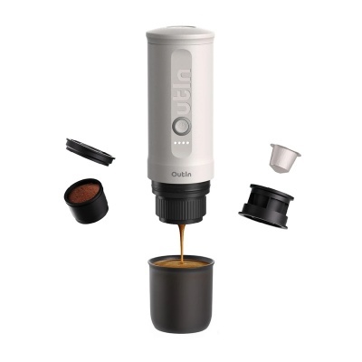 Máquina de café portátil a preparar café para um copo preto, com cápsulas e pó de café à volta