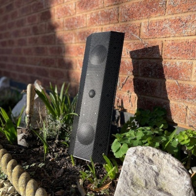 coluna preta de som com malha metálica no solo de jardim em frente a parede de tijolos vermelhos