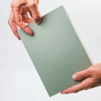 Caderno verde com a palavra ASK em relevo, segurado por duas mãos