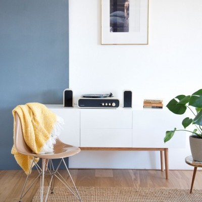 Sala com móvel branco e aparelho de som, cadeira transparente com manta amarela, planta em vaso e quadro na parede